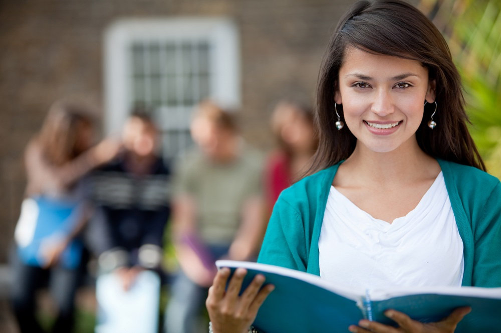 College aged female on a college campus reading a notebook and smiling. Peers behind her talking.