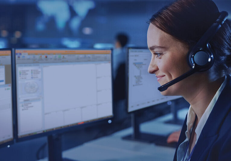 Woman working with a headset on. She is in a dark IT room working on multiple computer screens.