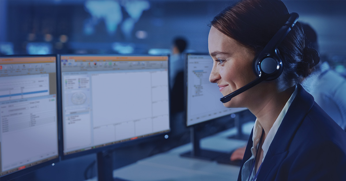 Woman working with a headset on. She is in a dark IT room working on multiple computer screens.