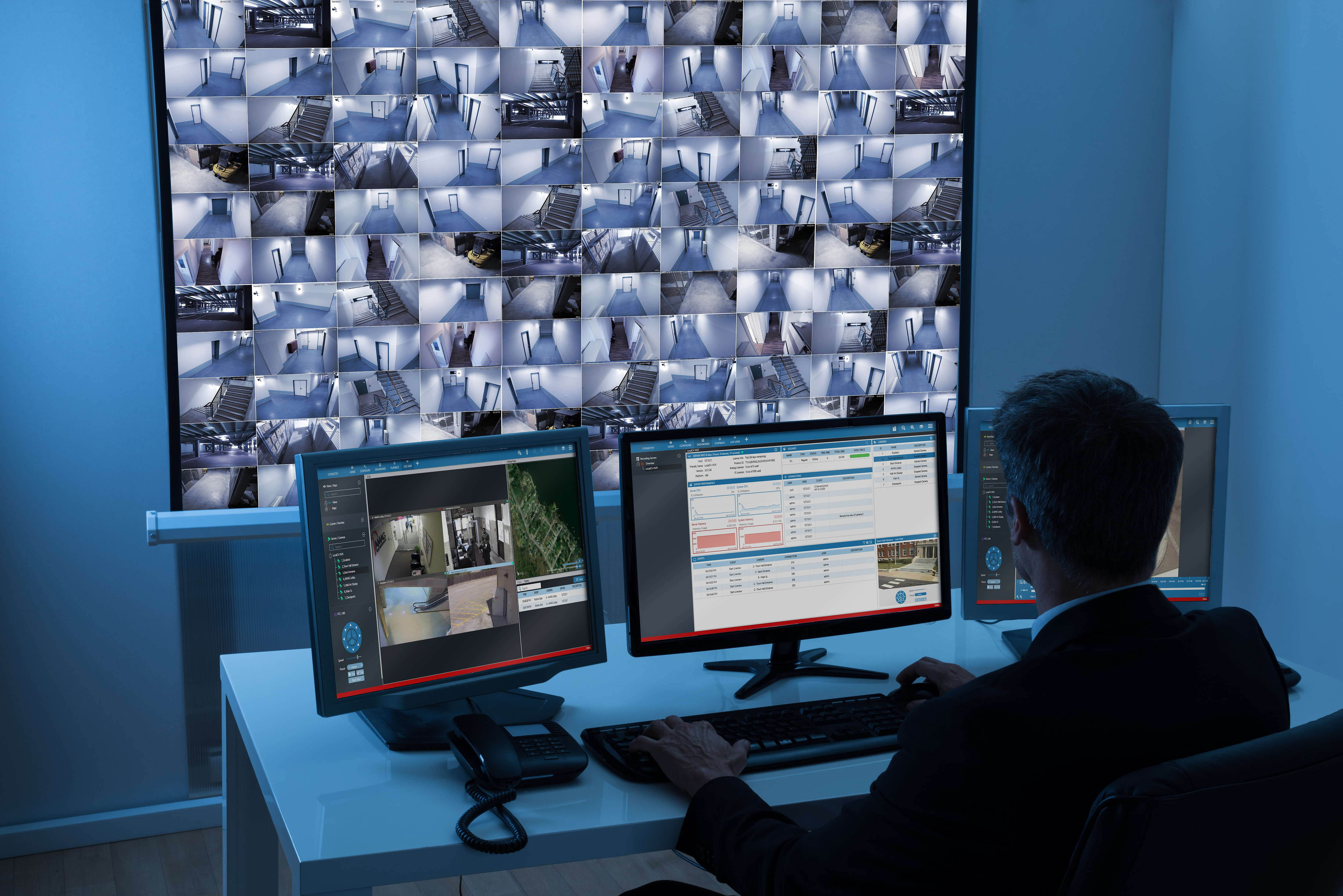 Man working in security room viewing security footage on large screen. He is also working on three computer screens. His back is to the camera.