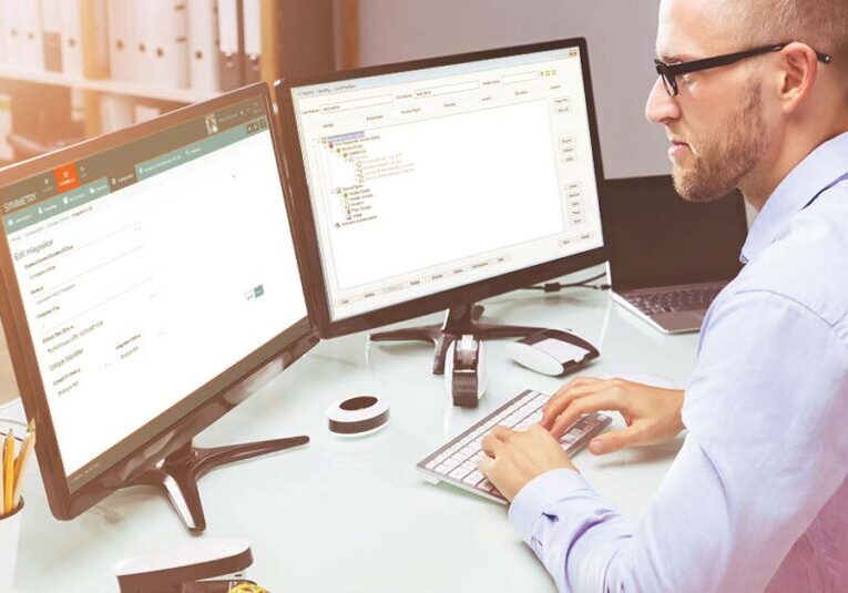 Man wearing glasses working at a desk. He is working on two computer screens.