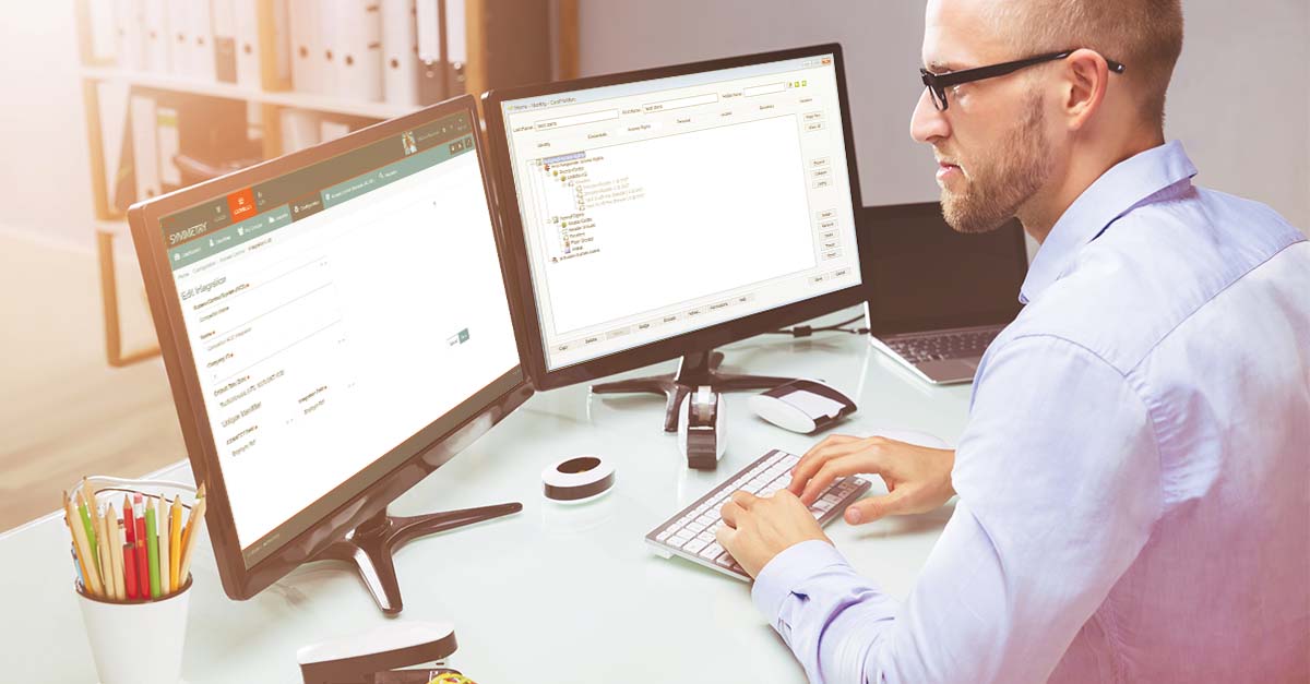 Man wearing glasses working at a desk. He is working on two computer screens.