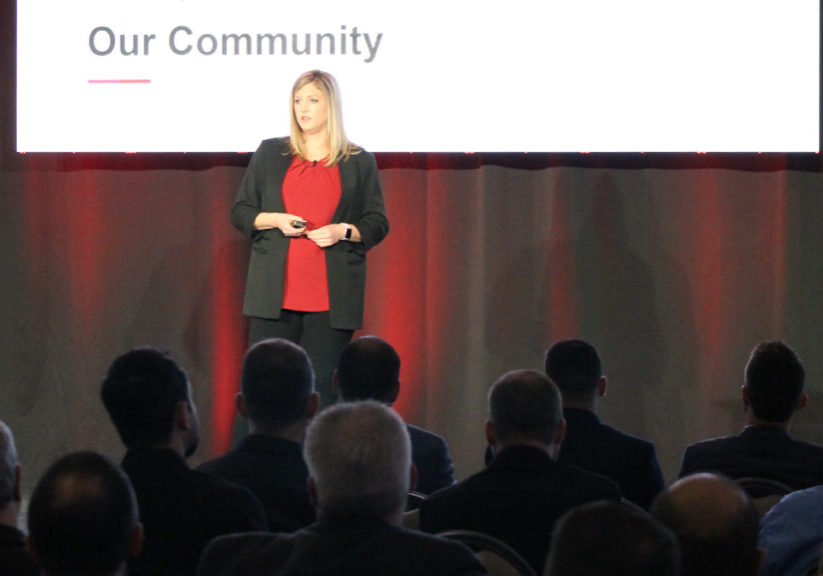 Photo of a female speaker on a stage at AMAG's annual Security Engineering Symposium. There is a screen behind her and a crowd listening.