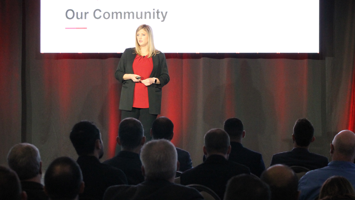 Photo of a female speaker on a stage at AMAG's annual Security Engineering Symposium. There is a screen behind her and a crowd listening.