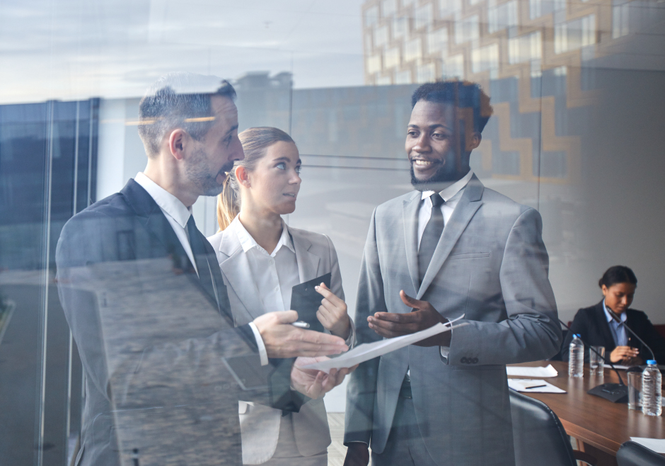 Group of three people discussing a paper in a conference room. There are two men and one woman. They are all wearing suits.